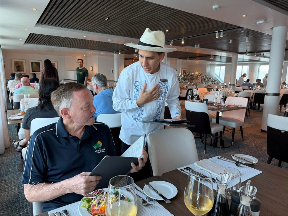 A diner seated at a restaurant table is reviewing the menu while a waiter in a traditional outfit is standing beside him, engaged in conversation. The restaurant has a modern interior with other diners in the background.