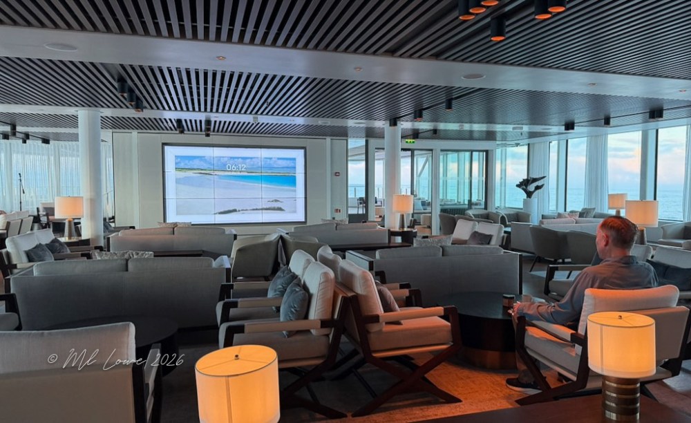 A modern lounge area with comfortable seating, large windows offering ocean views, and a screen displaying a timer and beach scenery. A person is seated, observing the screen.