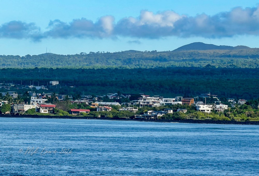 A coastal view showing residential buildings alongside a calm blue sea, with green hills and a blue sky in the background.