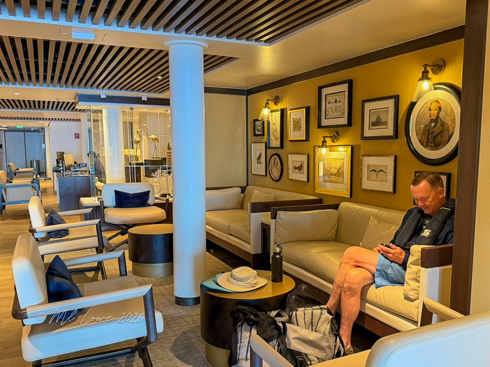 A cozy lounge area featuring modern seating, decorative wall art, and warm lighting. A man is seated on a couch, using his phone, while multiple chairs and a coffee table are arranged around him.