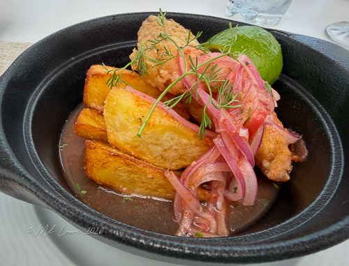 A serving of fried fish topped with pickled onions, alongside roasted plantains and a lime wedge, presented in a black dish.