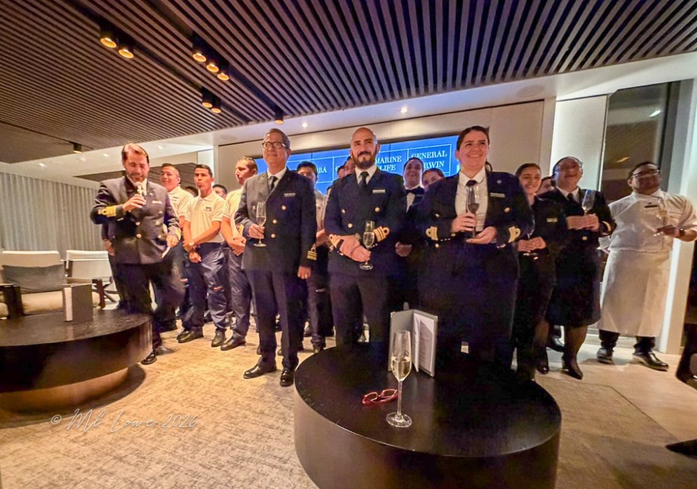 A group photo of ship crew members, including officers and staff, standing together in a modern indoor space, celebrating with drinks in hand.
