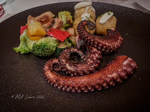 A beautifully plated dish featuring cooked octopus tentacles, served alongside a mix of colorful vegetables including broccoli, bell peppers, and mushrooms, on a dark round plate.