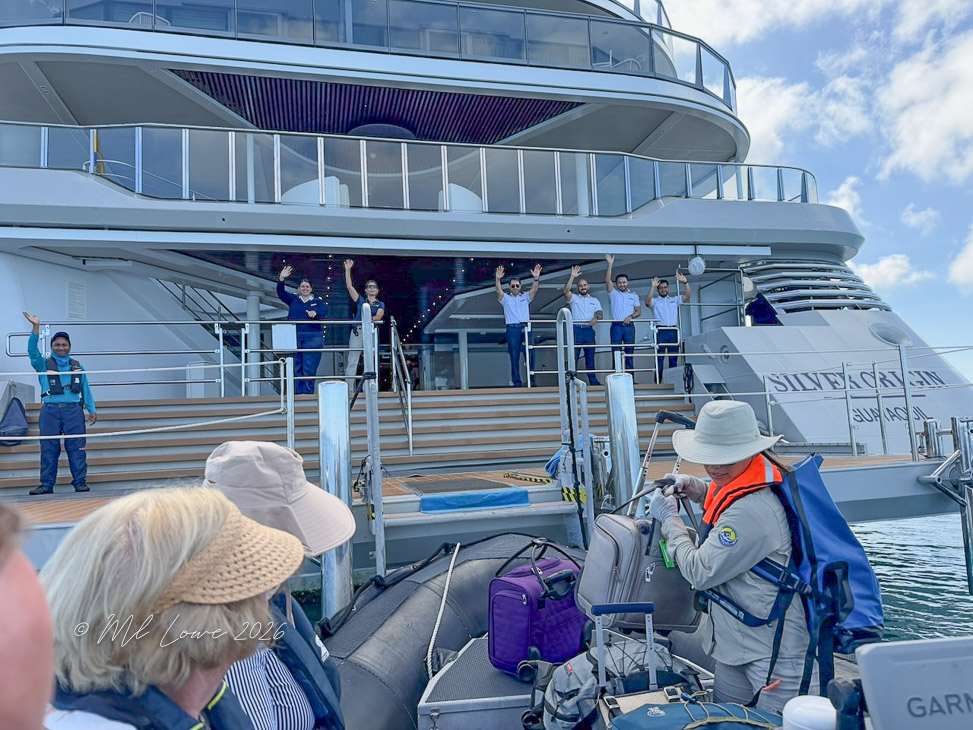 A group of crew members waves from a luxury cruise ship as passengers prepare to board a small inflatable boat nearby.