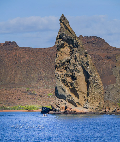 A tall, jagged rock formation rises from the sea, set against a backdrop of brown volcanic hills under a blue sky with scattered clouds. A small boat is visible near the shoreline.