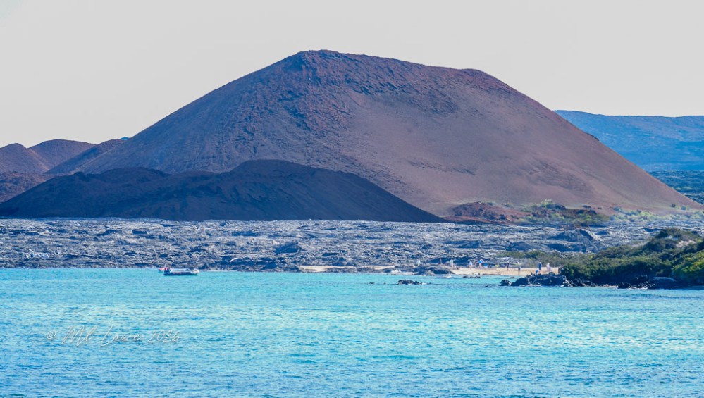 A scenic view of a brown volcanic mountain rising above a blue ocean, with rocky shoreline in the foreground and a small boat in the water.