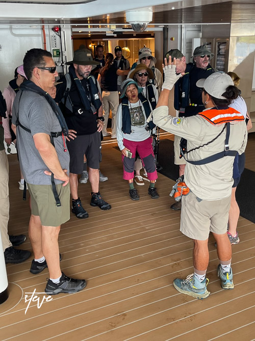 A group of people in casual attire with life jackets gathered on a ship's deck, listening to a guide who is explaining something. Various hats and sunglasses are visible.